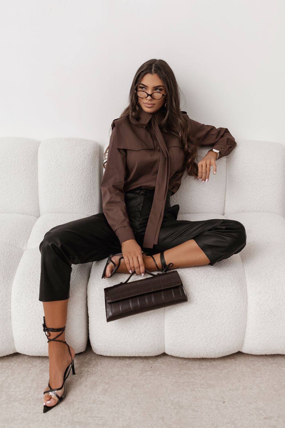 Woman in brown top and black pants sitting on a white couch holding a brown handbag.