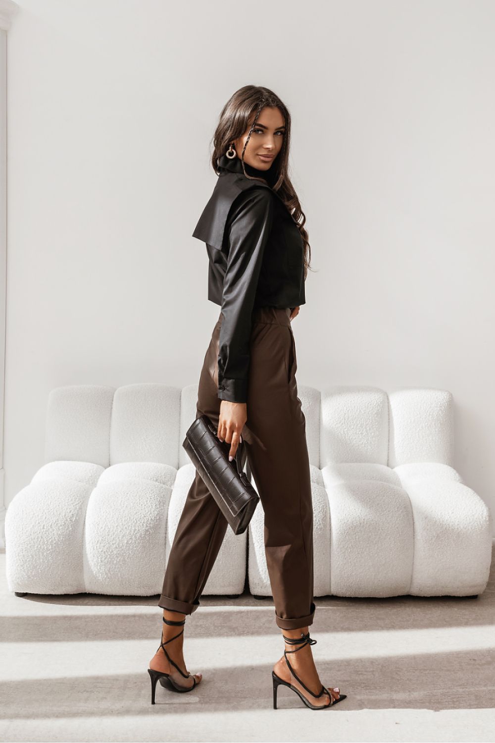 Woman in black leather jacket and brown pants holding a clutch in a modern interior setting.