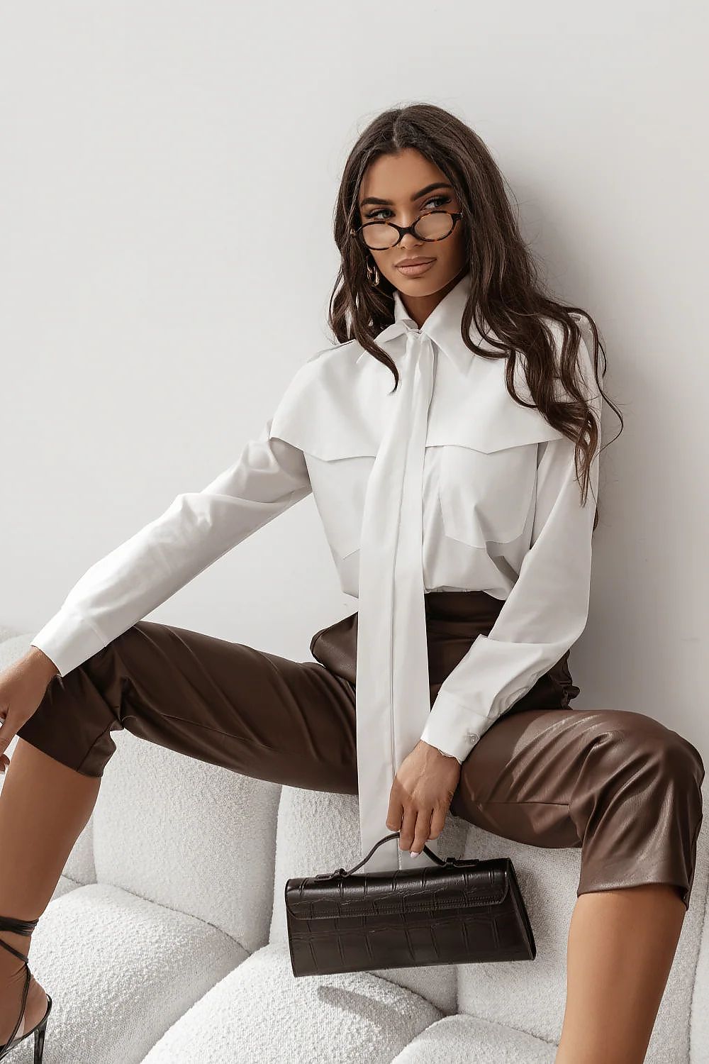 Woman wearing a white blouse with a tie and brown pants, sitting on a white couch.
