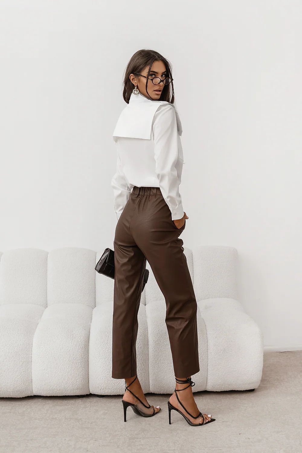 Woman wearing a white blouse and brown pants in a minimalistic room.