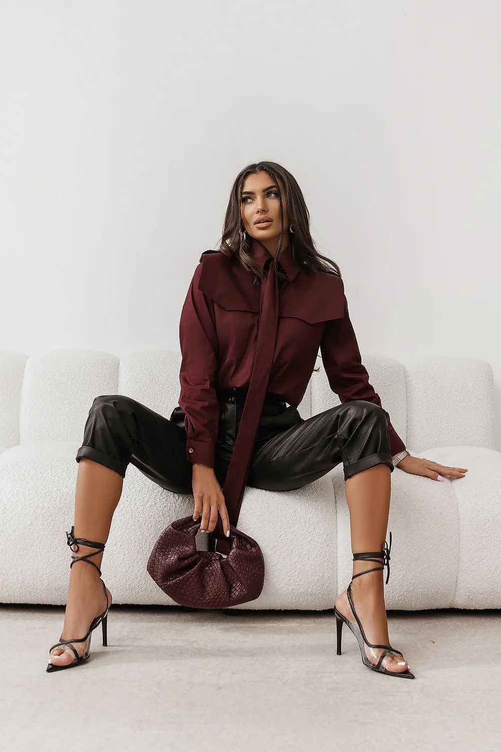 Woman in burgundy shirt and black pants sitting on a white couch holding a brown handbag.