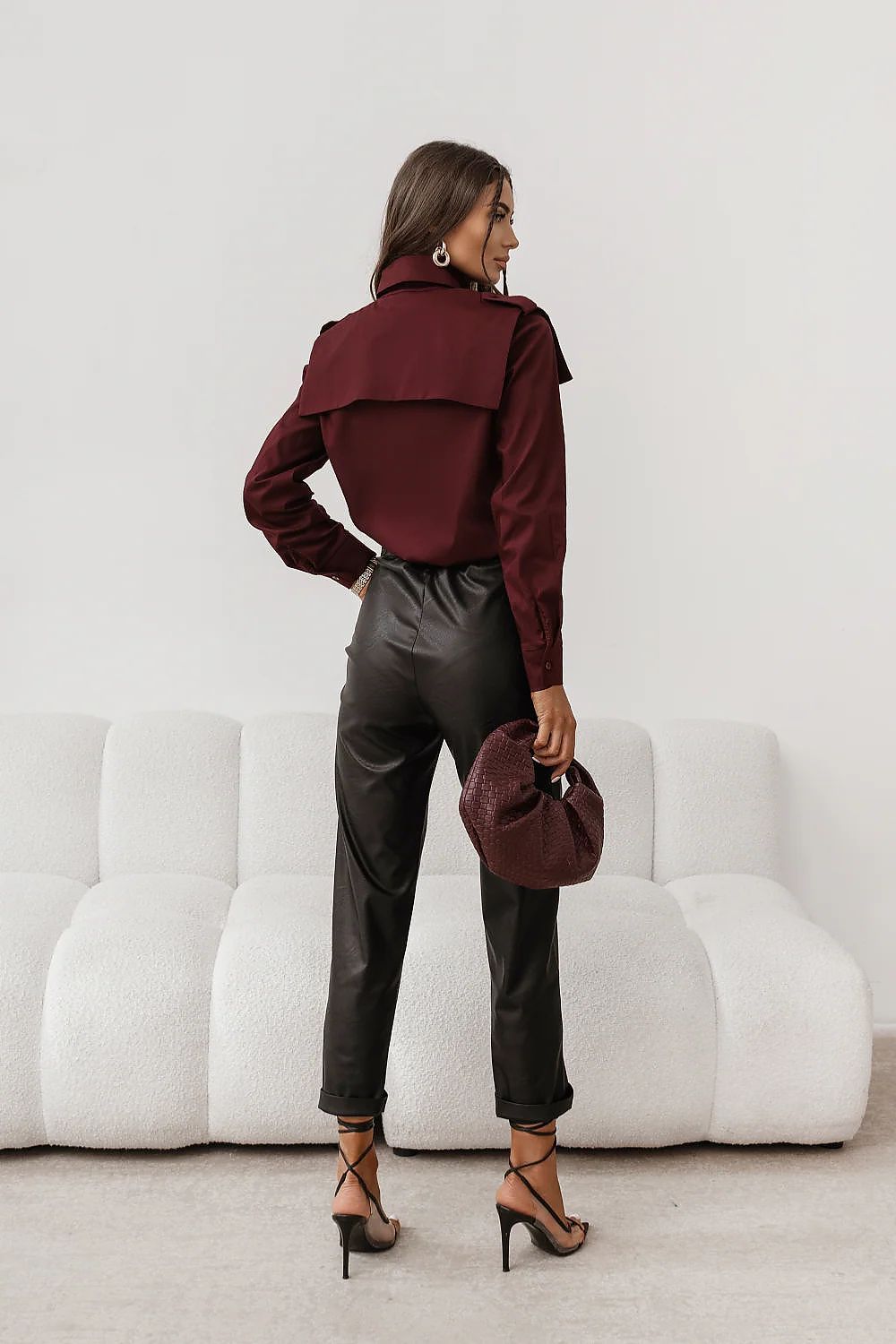 Woman in burgundy shirt and black pants holding a matching hat in a minimalistic setting.