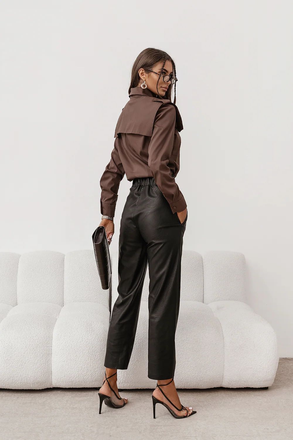 Woman in a brown blouse and black pants holding a clutch in a minimalistic setting.