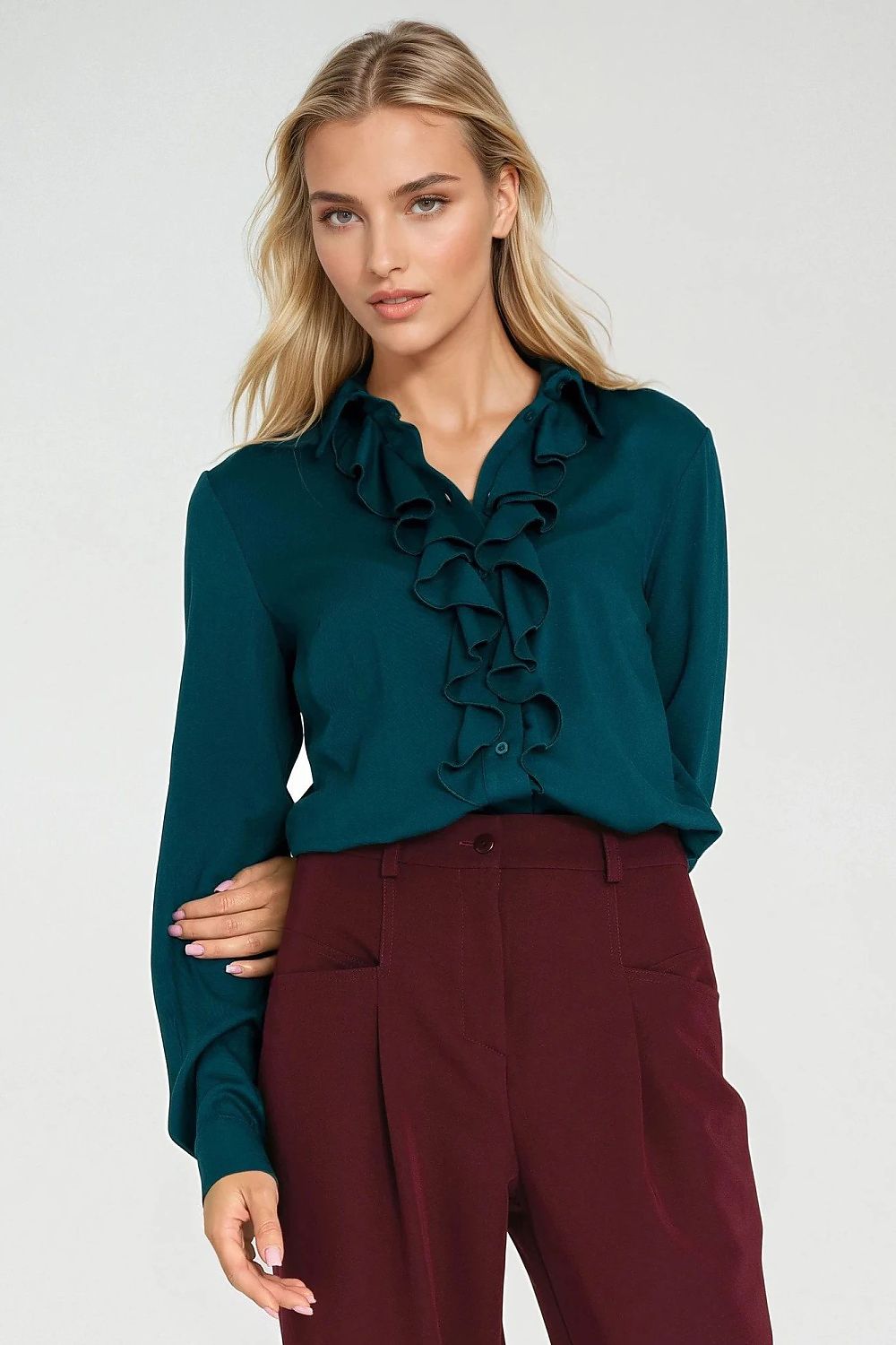 Woman wearing a teal blouse with ruffle details and burgundy pants on a white background