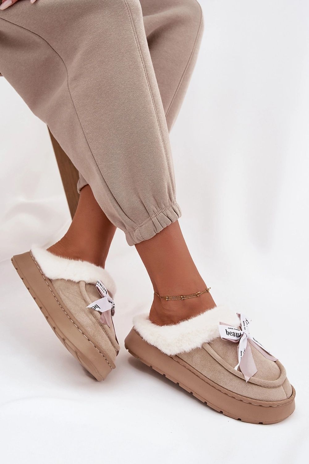 Beige slippers with white fur trim worn with beige pants on a light background