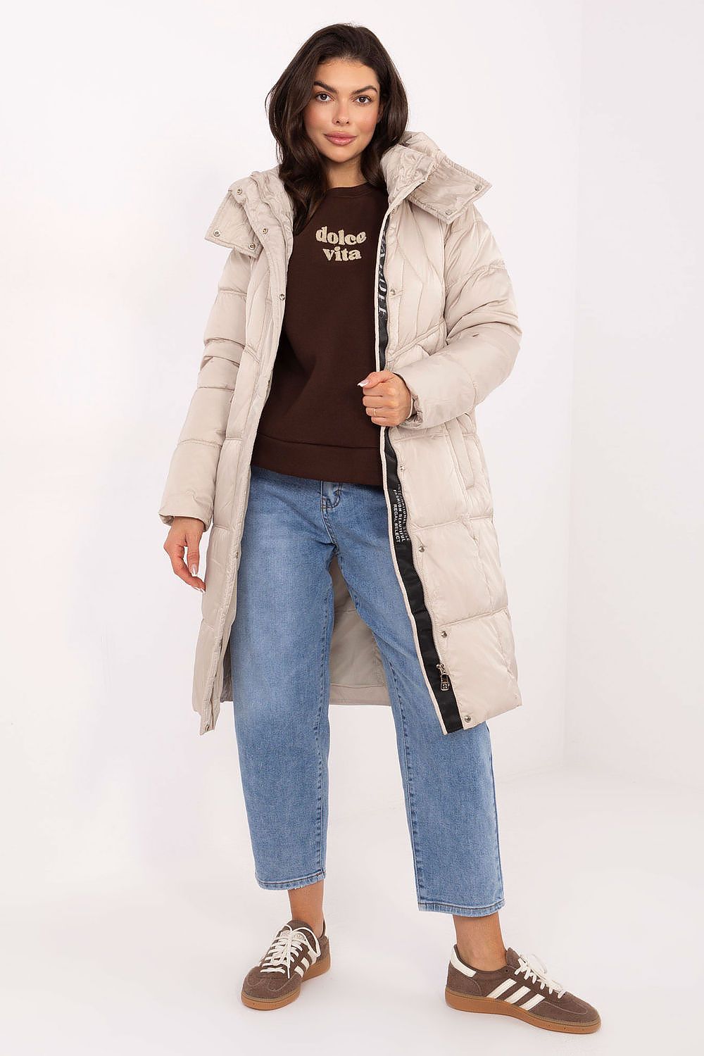 Woman wearing a beige puffer coat, brown t-shirt, blue jeans, and brown sneakers on a white background