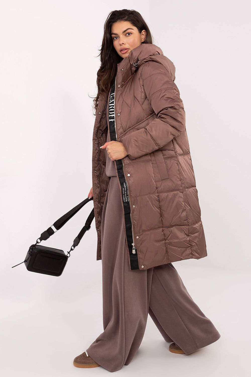 Woman wearing a long brown puffer coat with a black handbag on a white background