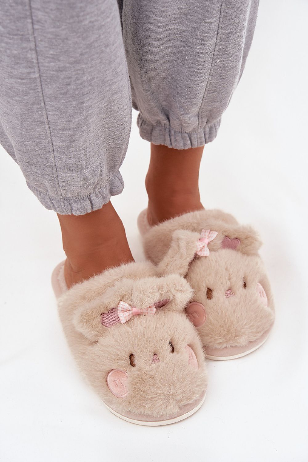 Beige slippers with pink bows worn by a person in gray pants on a white background