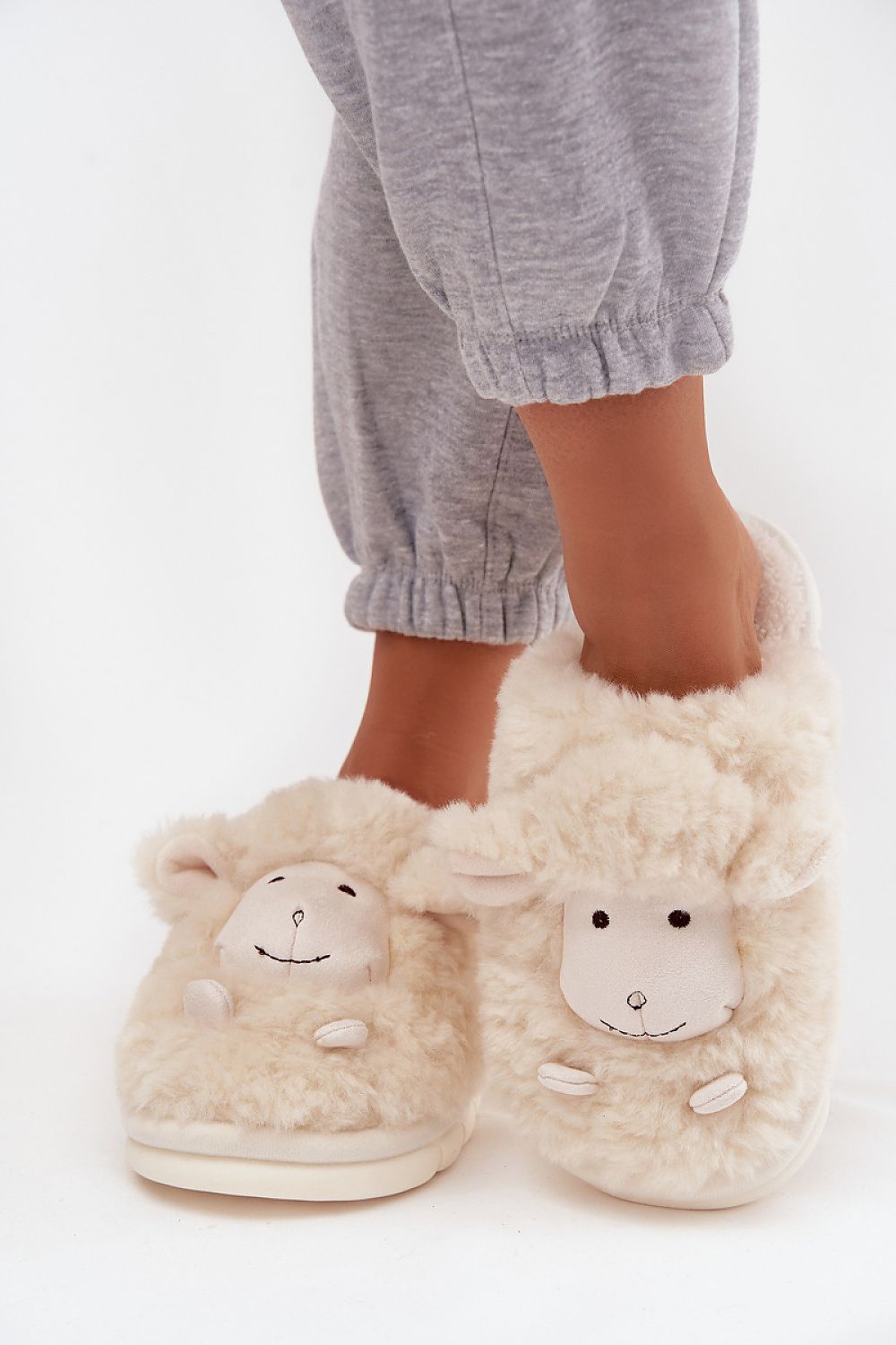 Beige slippers with sheep faces worn by a person in gray pants on a white background