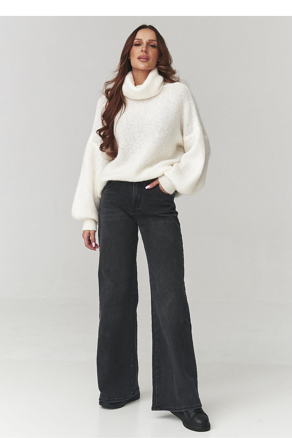 Woman wearing a white sweater and black jeans on a plain background