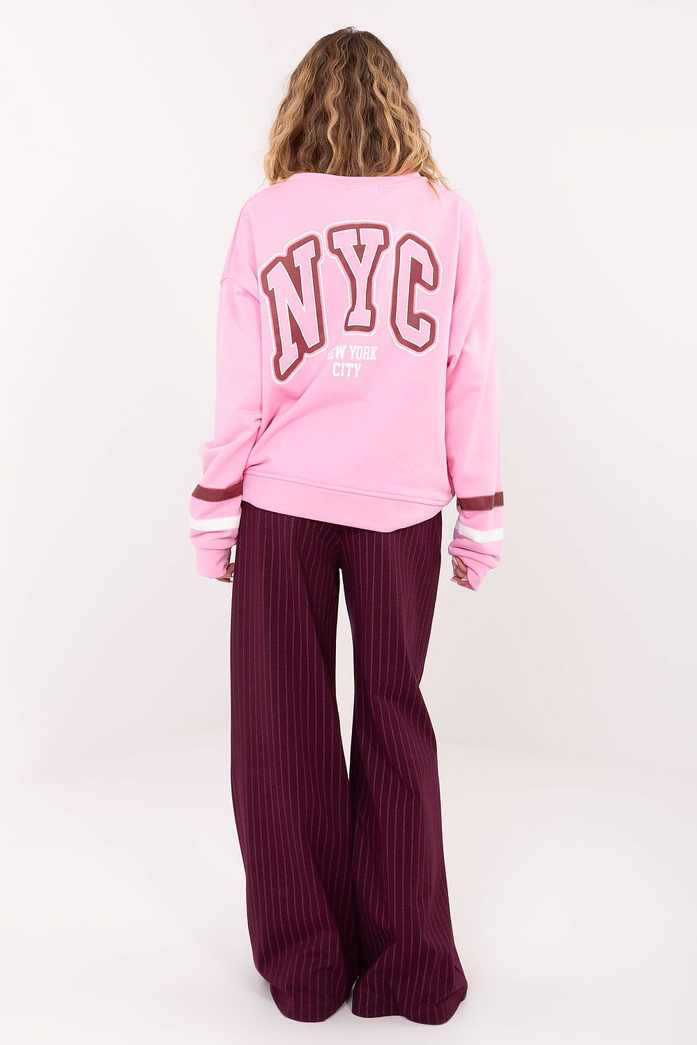 Person wearing a pink sweatshirt with 'NYC' text and purple pants on a white background