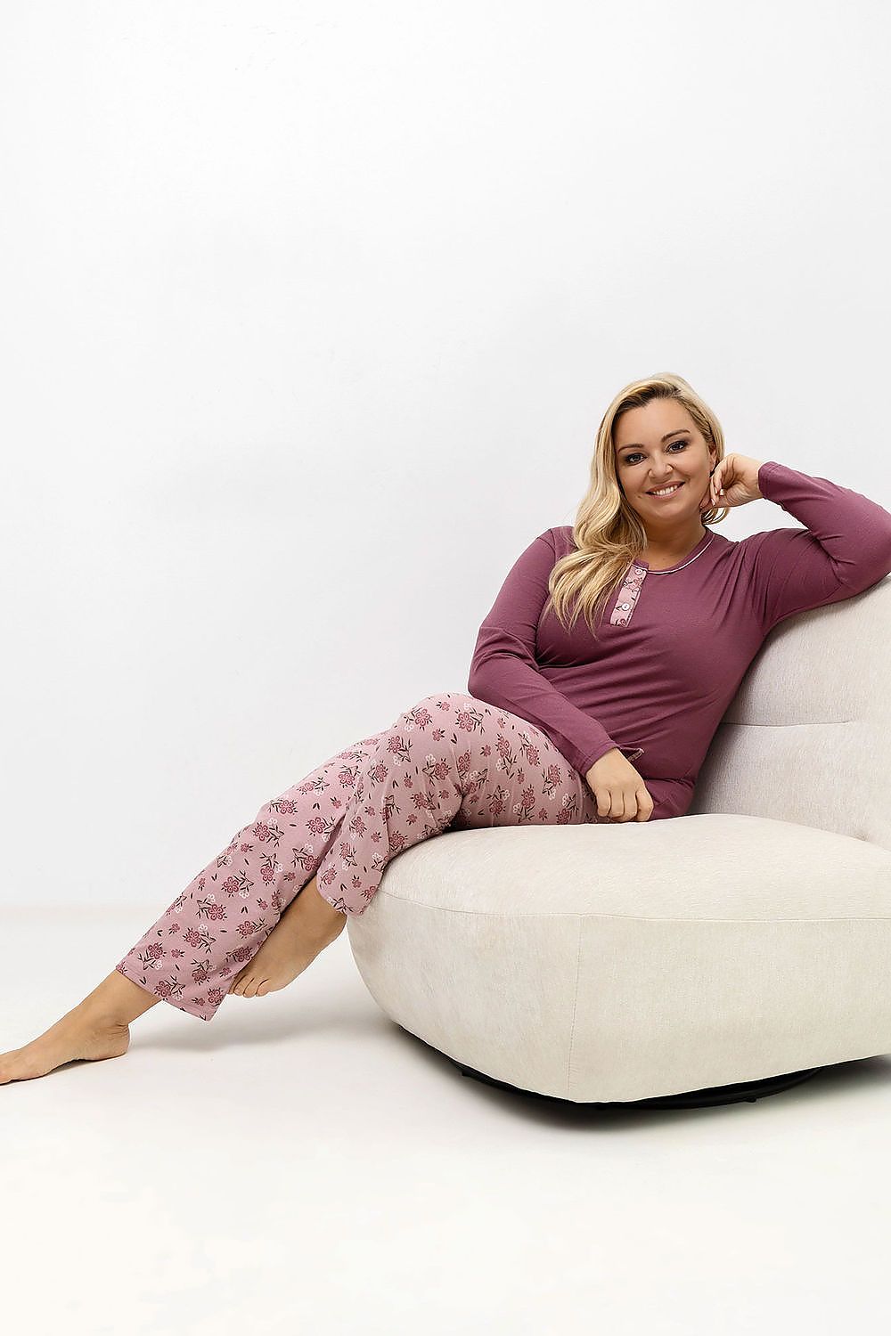 Woman wearing a purple top and pink floral pants sitting on a white chair against a white background