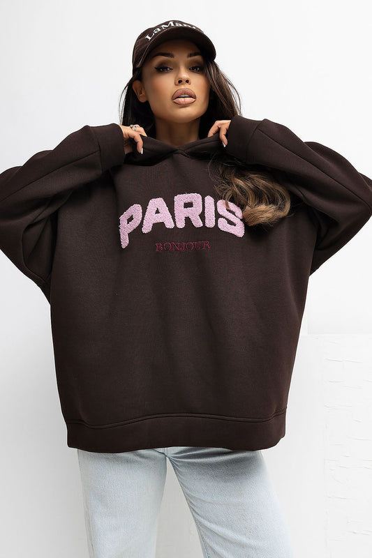 Person wearing a brown hoodie with 'PARIS' text, light blue jeans, and a cap on a white background