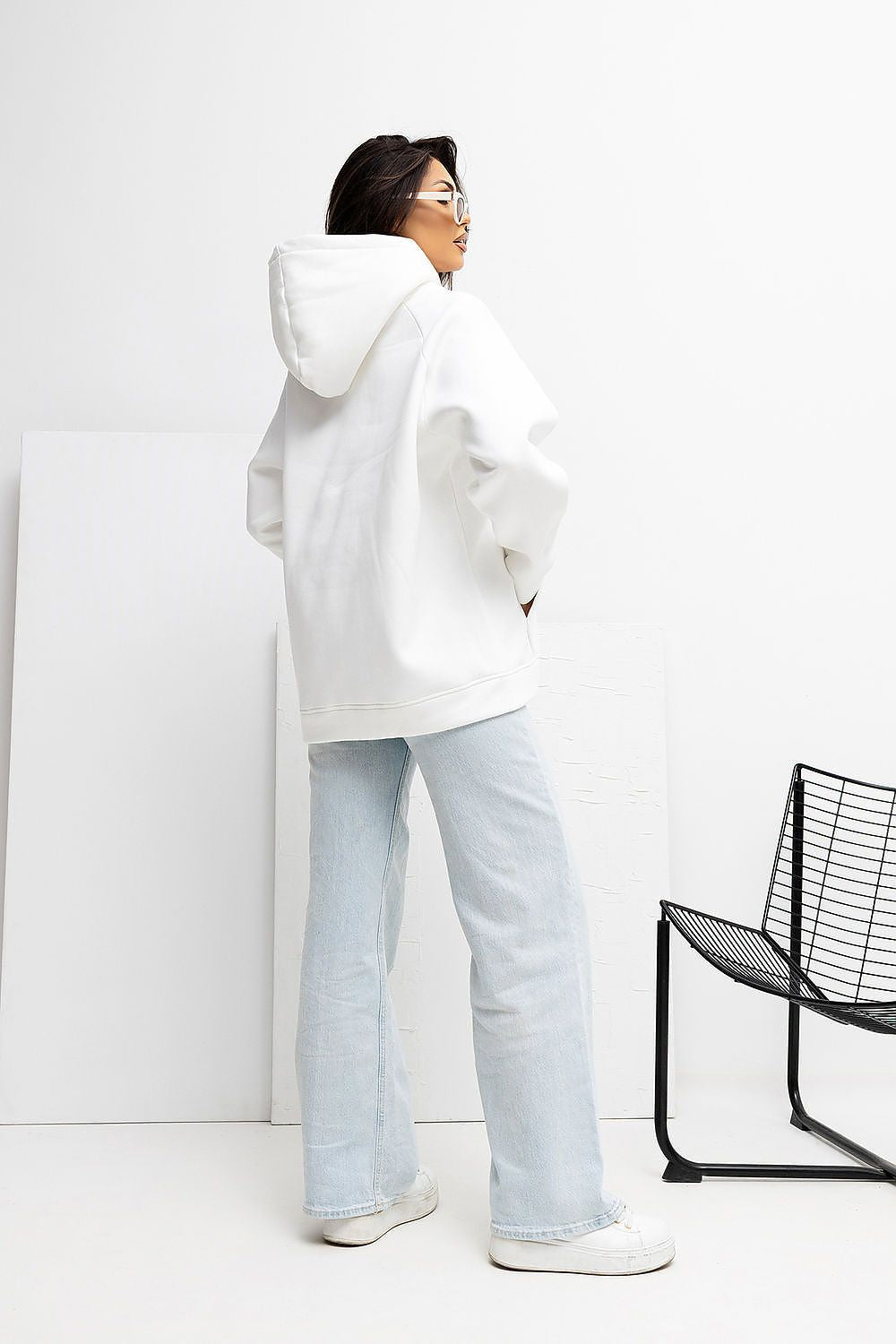 Person wearing a white hoodie and light blue jeans standing against a white background with a black chair.