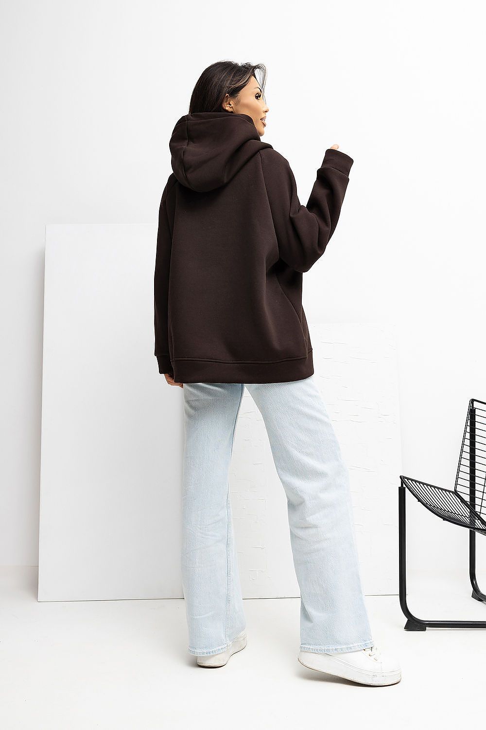 Person wearing a brown hoodie and light blue jeans standing against a white wall with a black chair.