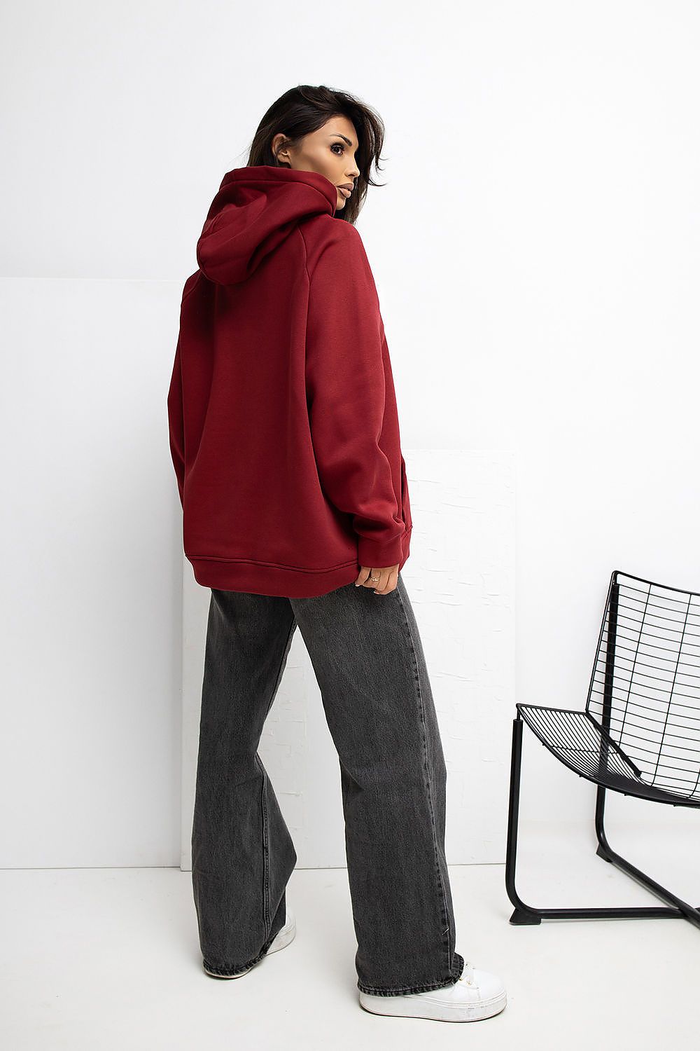 Person wearing a red hoodie and gray pants standing against a white wall with a black chair.