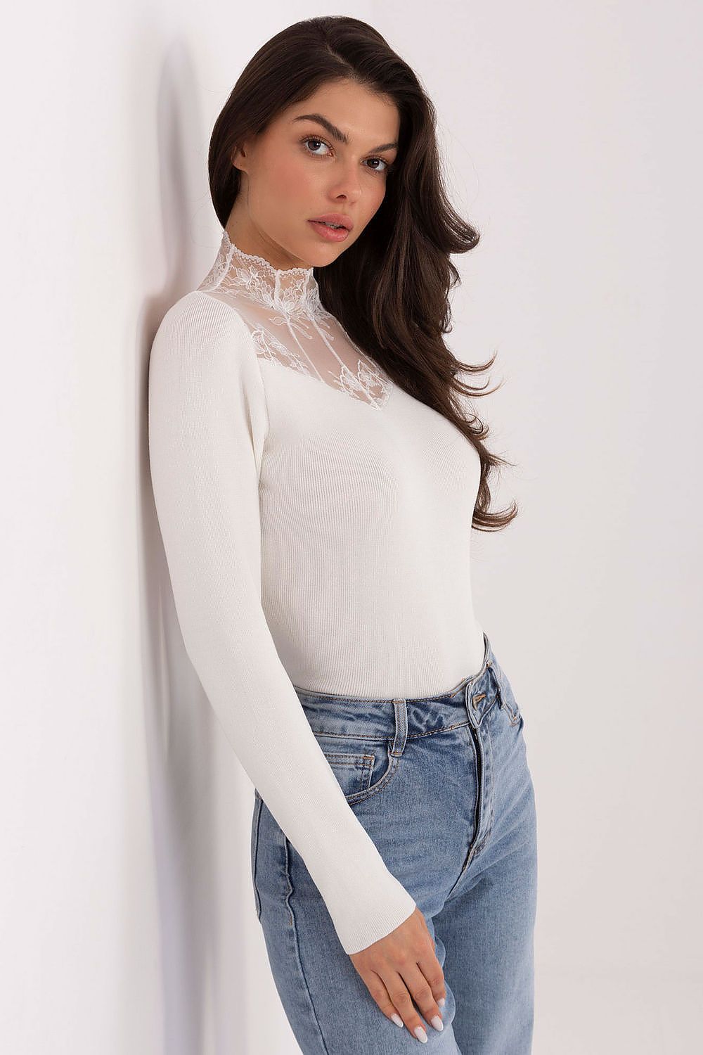 Woman wearing a white long-sleeve top with lace detailing and blue jeans against a white background