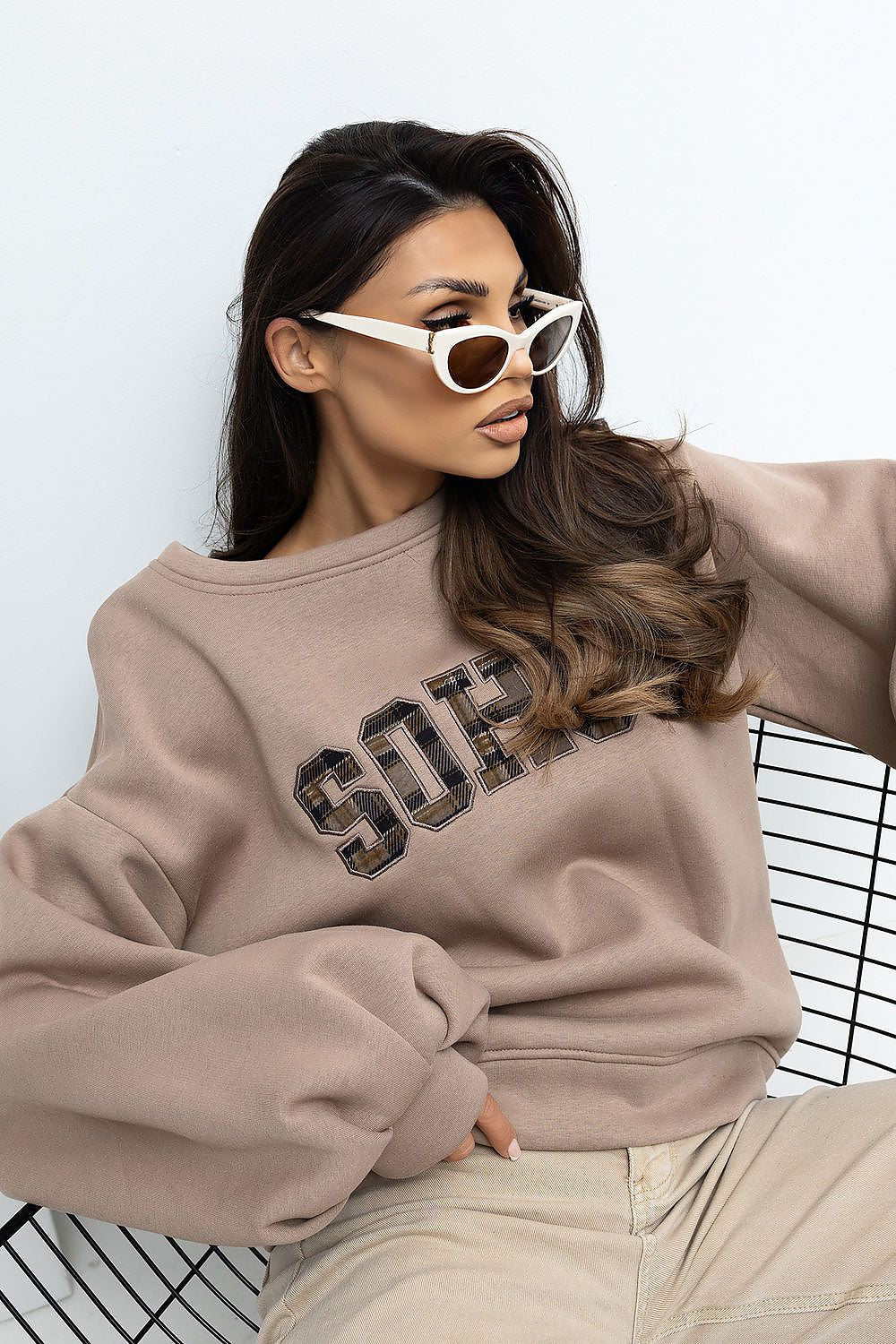 Woman wearing a beige sweatshirt with 'SOPH' text, white sunglasses, and beige pants on a light background