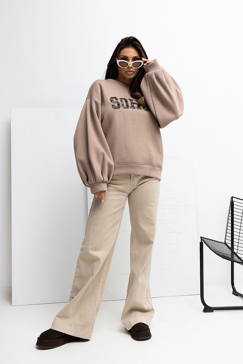 Woman wearing a beige oversized sweatshirt and pants with sunglasses indoors.