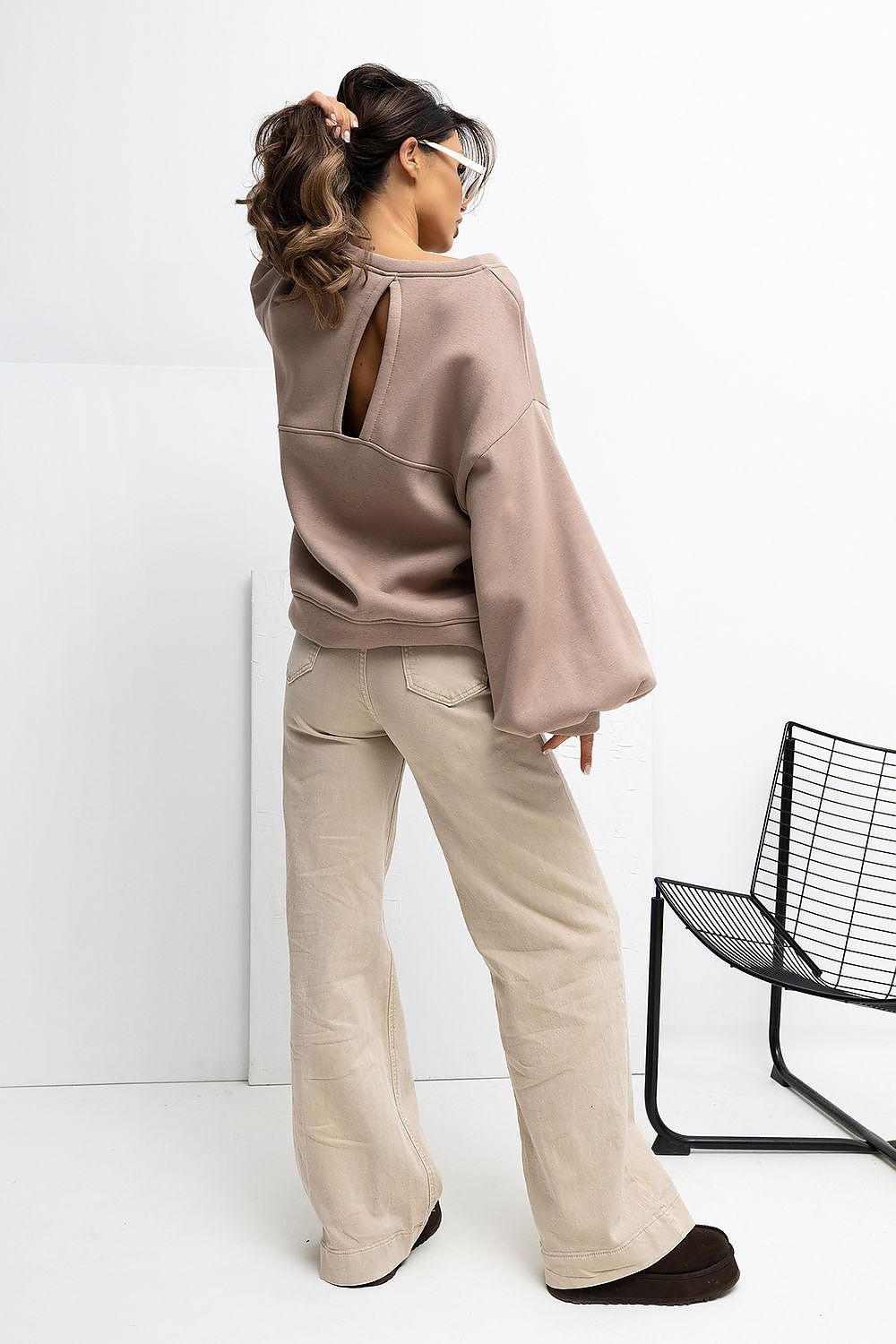 Person wearing a beige sweater and pants in a minimalistic setting with a black chair.