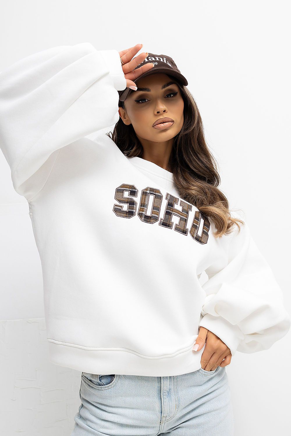 Woman wearing a white sweatshirt with 'SOHO' text, light blue jeans, and a cap on a white background