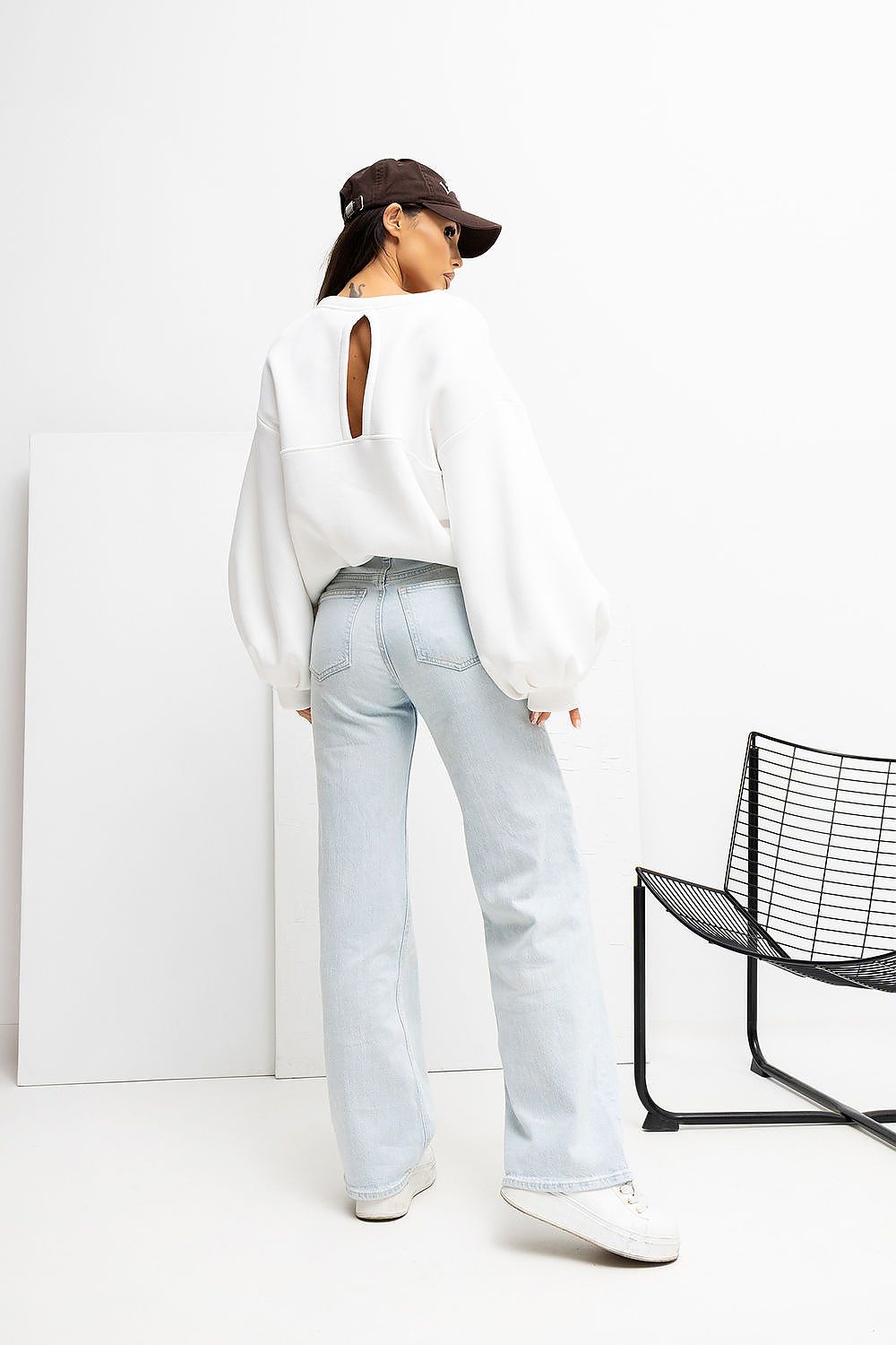 Person wearing a white sweater, light blue jeans, and a black cap in a minimalistic setting.