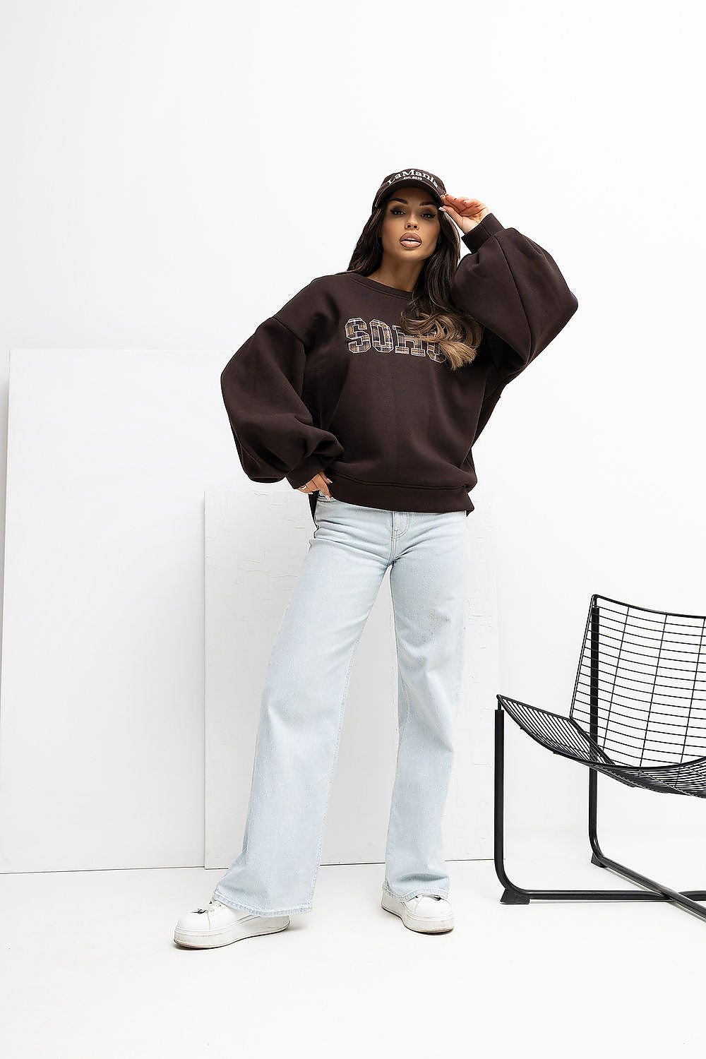 Person wearing a dark brown sweatshirt and light blue jeans standing against a white wall with a black chair.