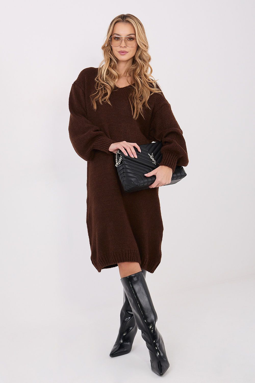 Woman wearing a brown dress and black boots holding a black clutch against a white background