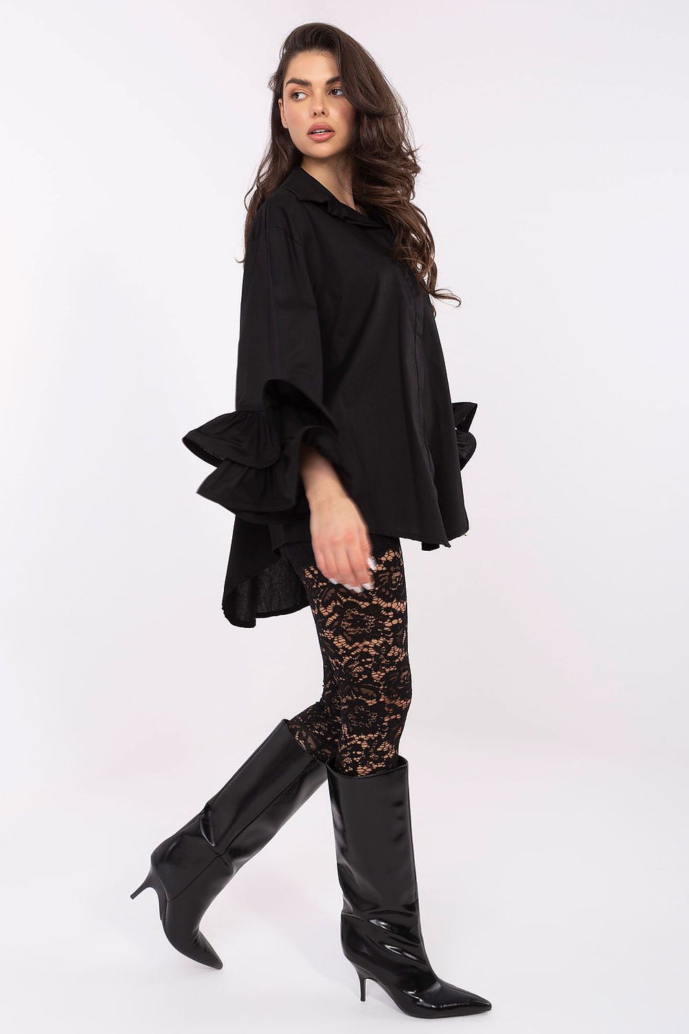 Woman wearing a black outfit with a ruffled top, patterned leggings, and black knee-high boots on a white background.
