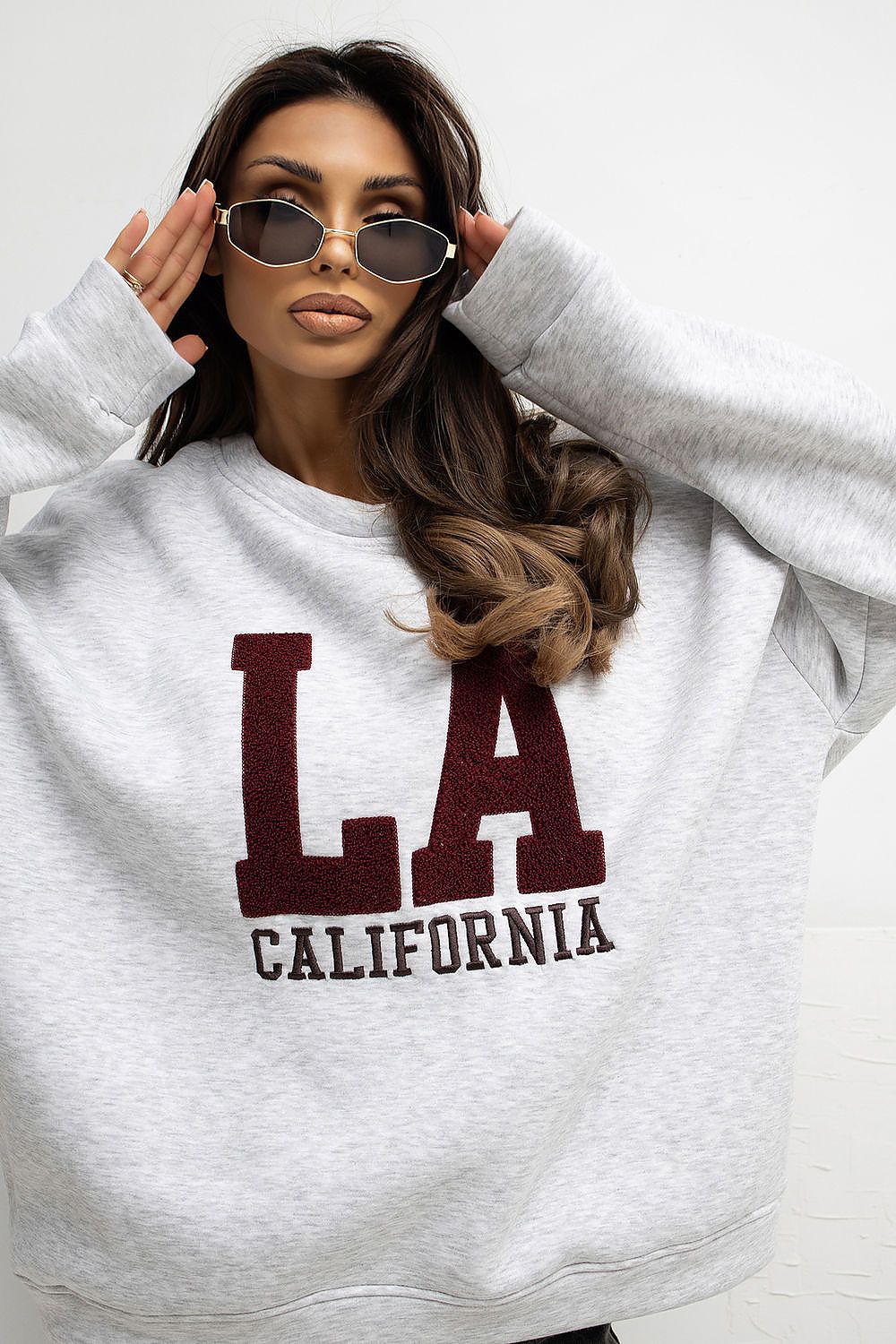 Woman wearing a gray sweatshirt with 'LA California' text, holding sunglasses.