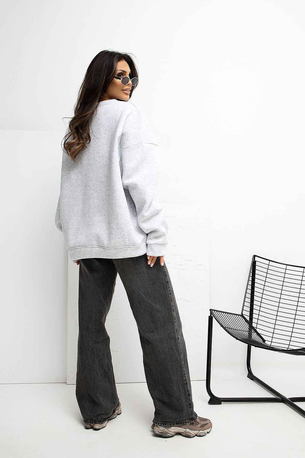 Person wearing a gray oversized sweatshirt and dark jeans standing against a white wall with a black chair.