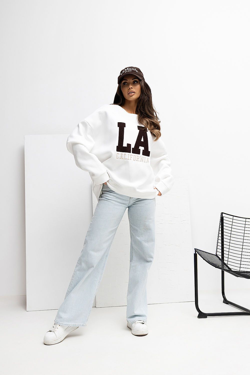 Person wearing a white sweatshirt with 'LA' letters, light blue jeans, and a cap in a minimalistic setting.