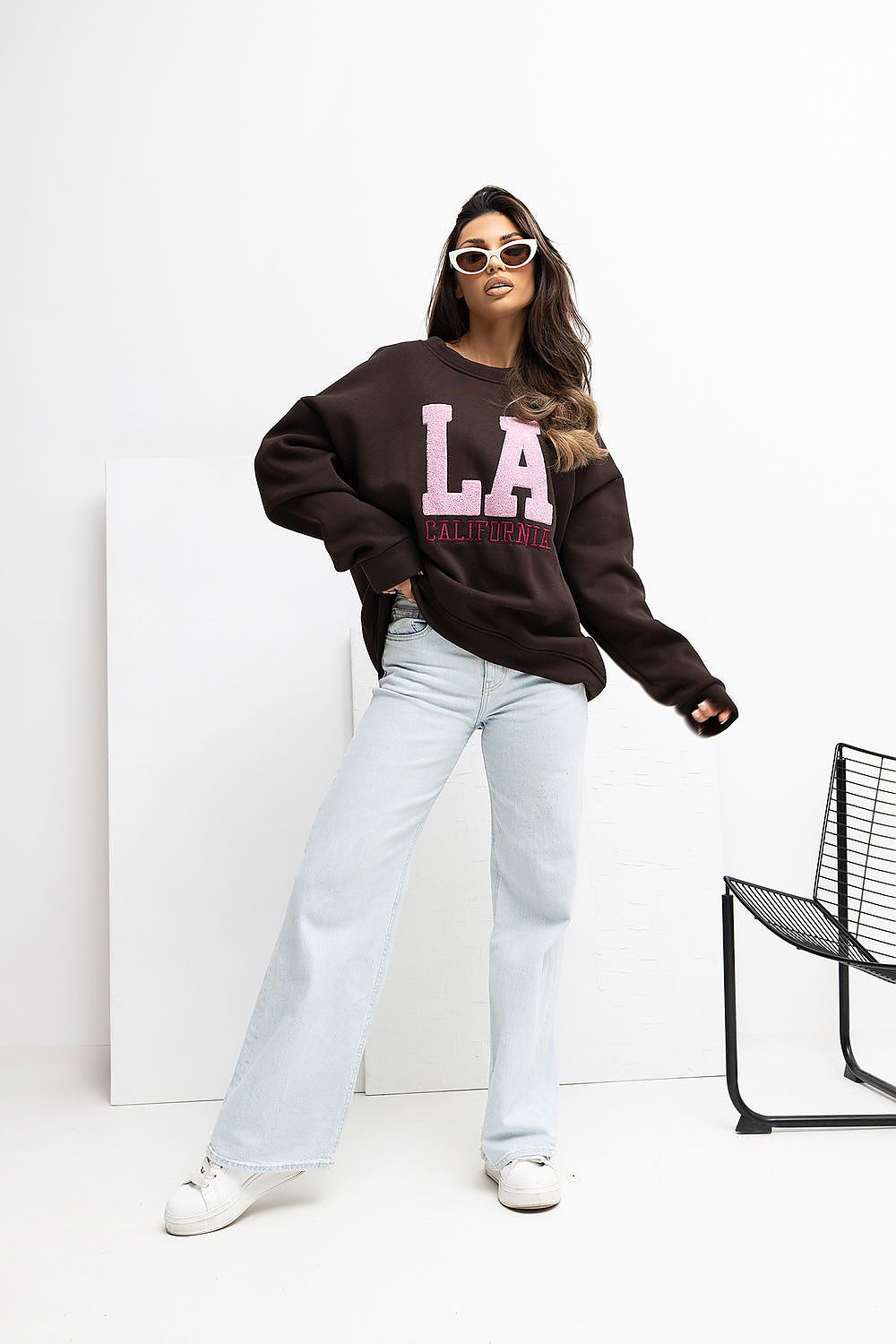 Person wearing a brown sweatshirt with 'LA California' text, light blue jeans, and white sneakers on a white background.