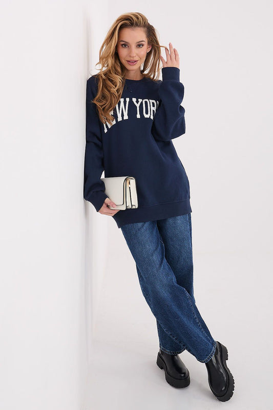 Woman wearing a navy blue 'New York' sweatshirt and jeans, holding a white clutch against a white background.