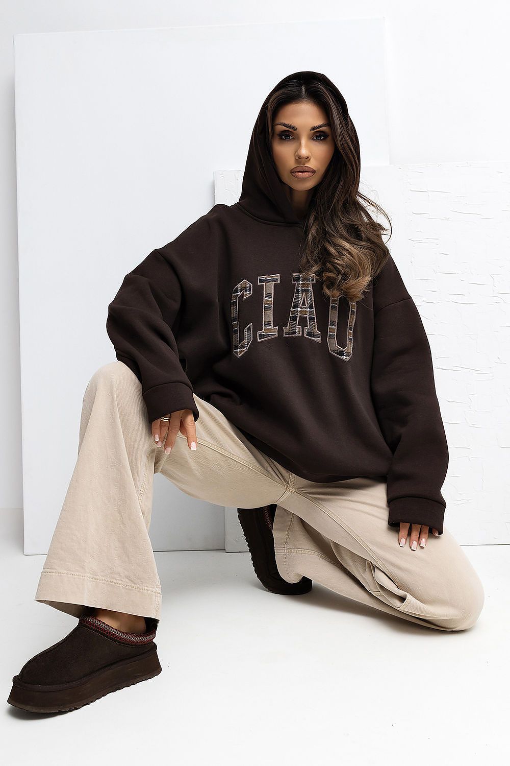 Woman wearing a brown 'CIAO' sweatshirt and beige pants sitting on the floor against a white wall.