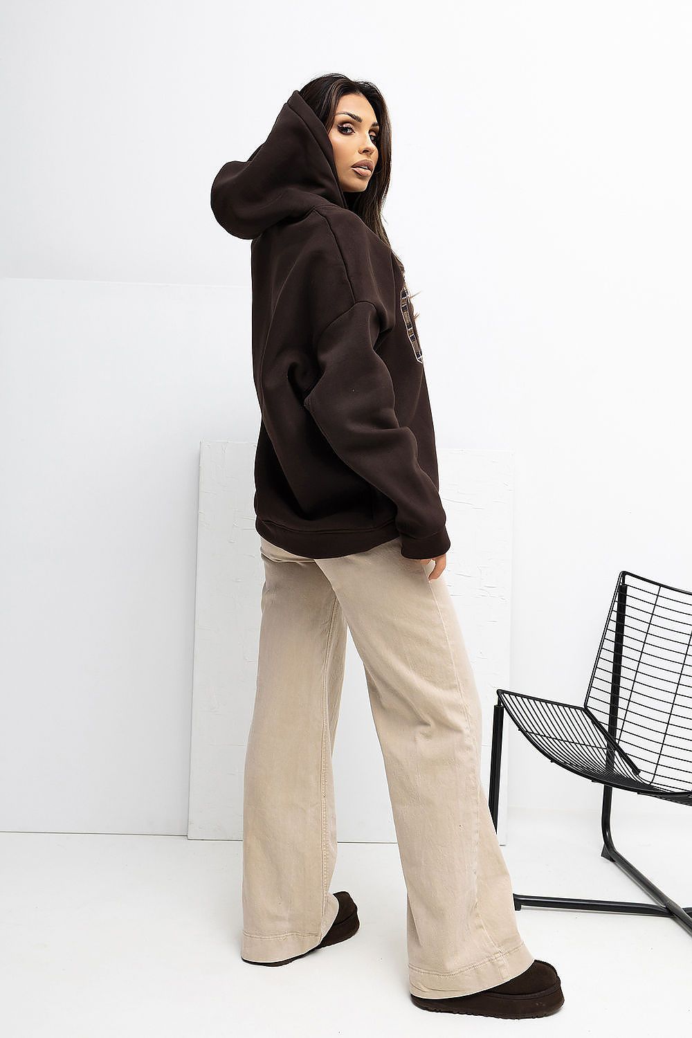 Person wearing a brown hoodie and beige pants standing against a white wall with a black chair.