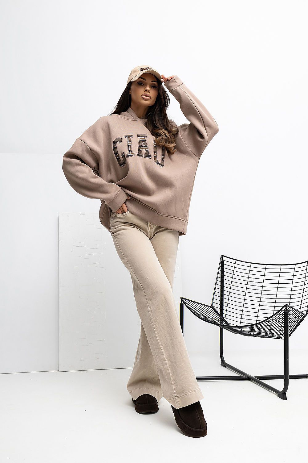 Person wearing a beige oversized sweatshirt with text, beige pants, and black shoes in a minimalistic setting.