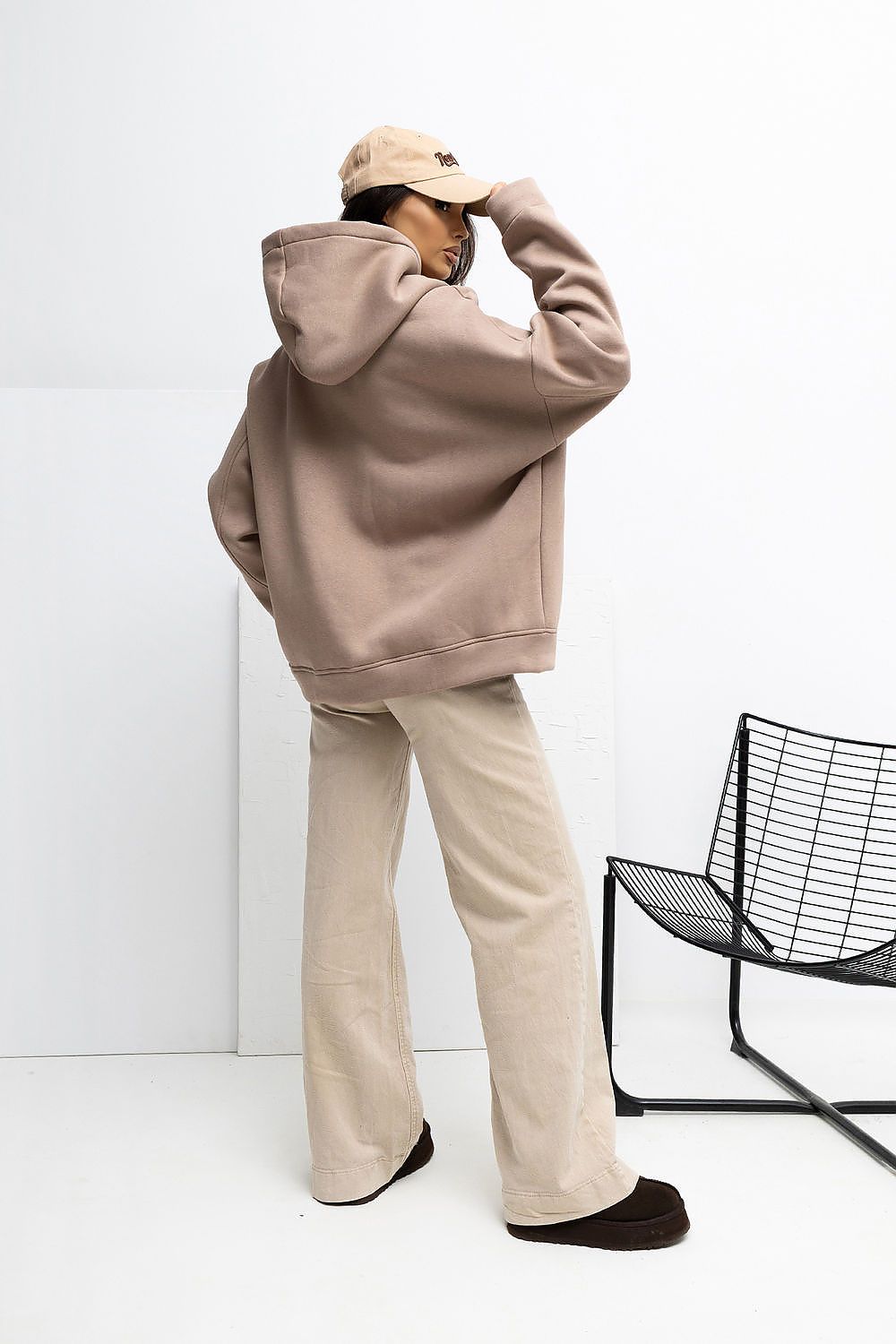 Person wearing a beige hoodie and pants with a cap, standing against a white background with a black chair.