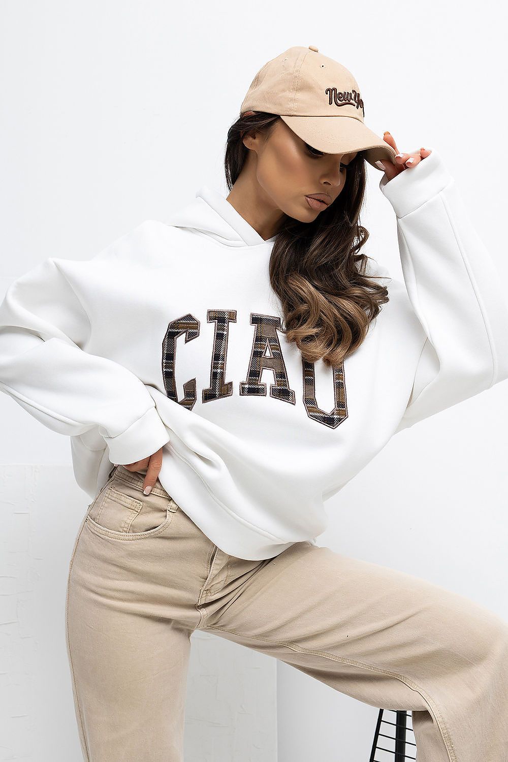 Person wearing a white hoodie with text, beige cap, and pants on a white background