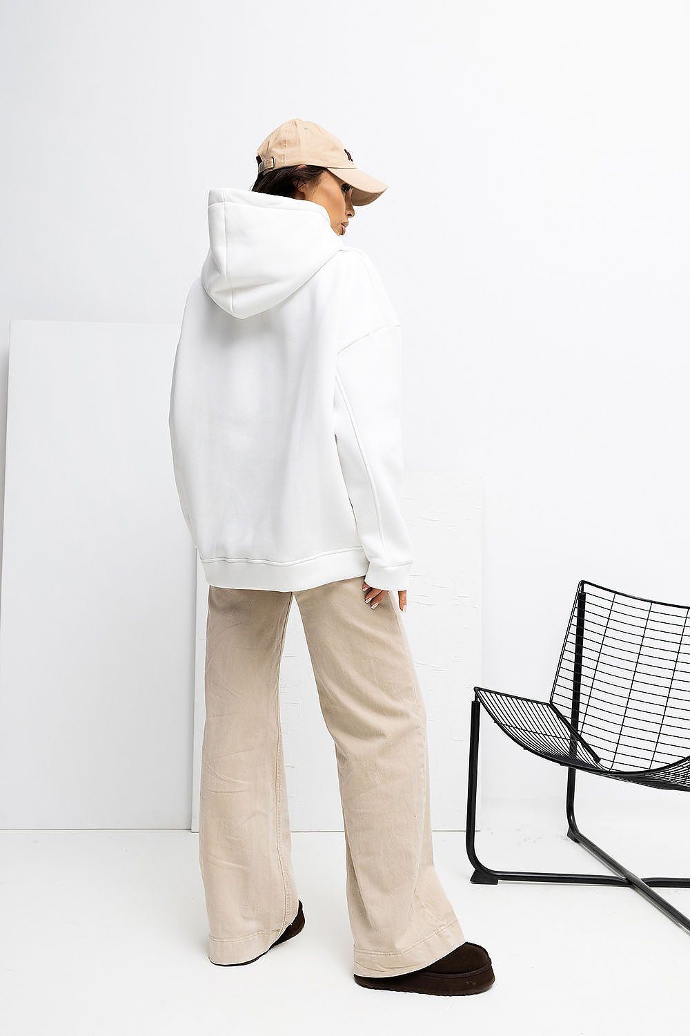 Person wearing a white hoodie and beige pants standing next to a black chair against a white wall.