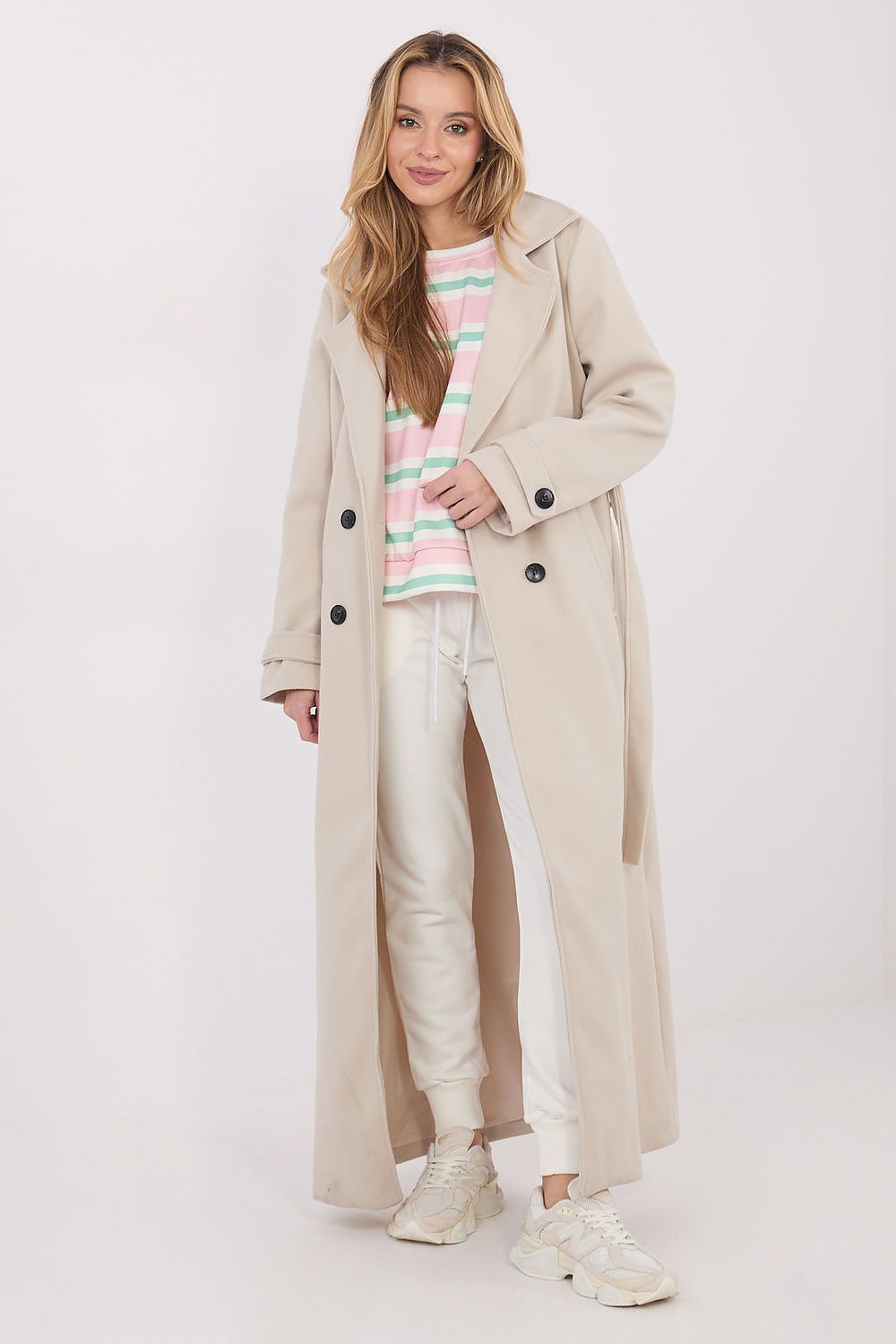 Woman wearing a beige trench coat over a colorful striped sweater and white pants on a white background