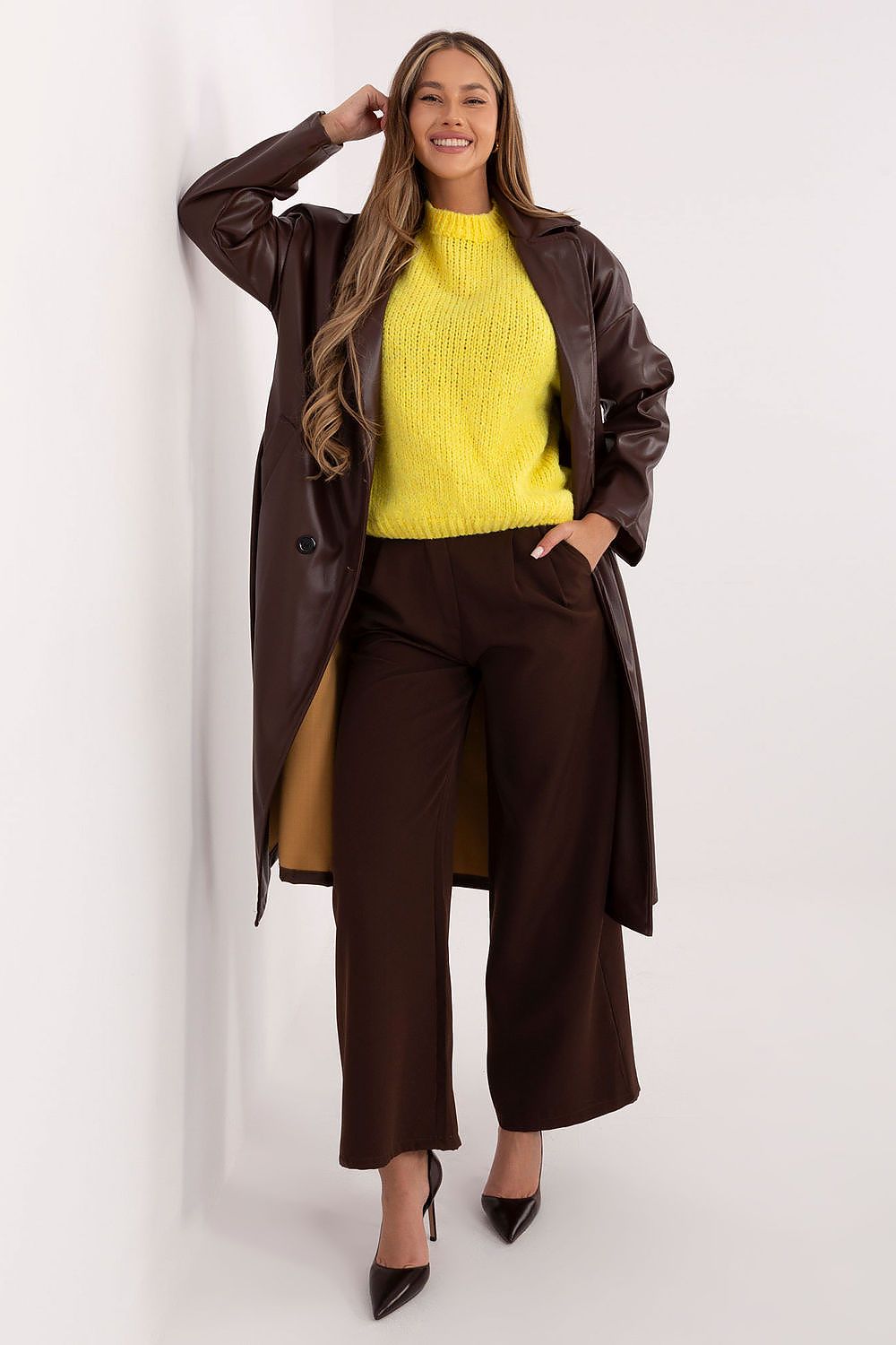 Woman wearing a yellow sweater and brown pants with a long brown coat against a white background