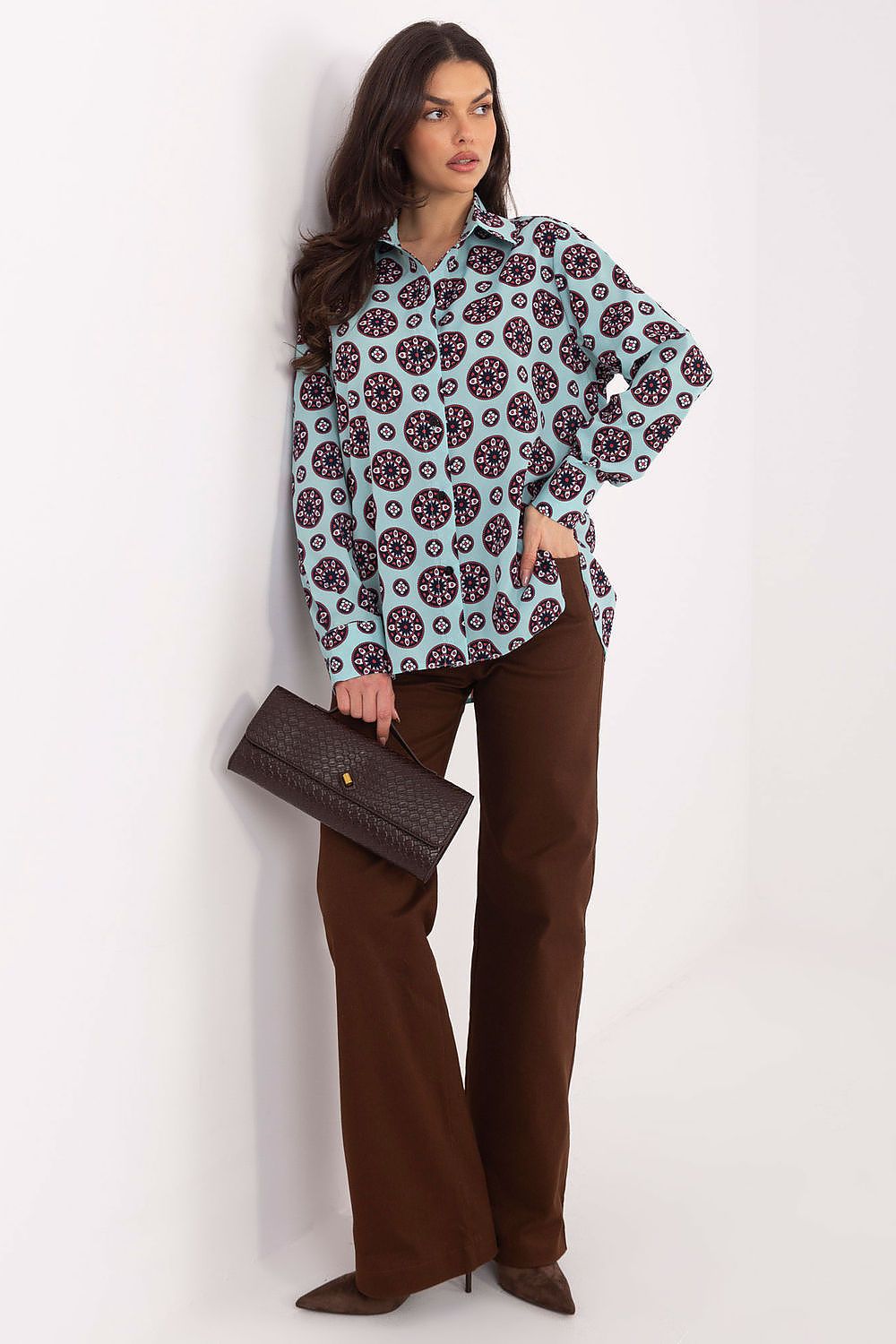 Woman wearing a patterned blouse and brown pants holding a clutch against a white background