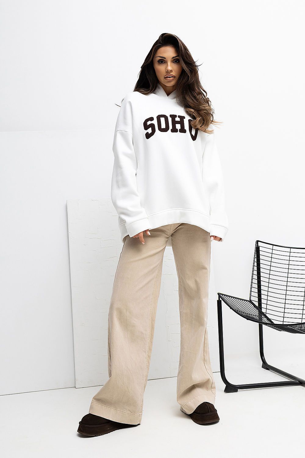 Woman wearing a white hoodie with 'SOHO' text and beige pants against a white background