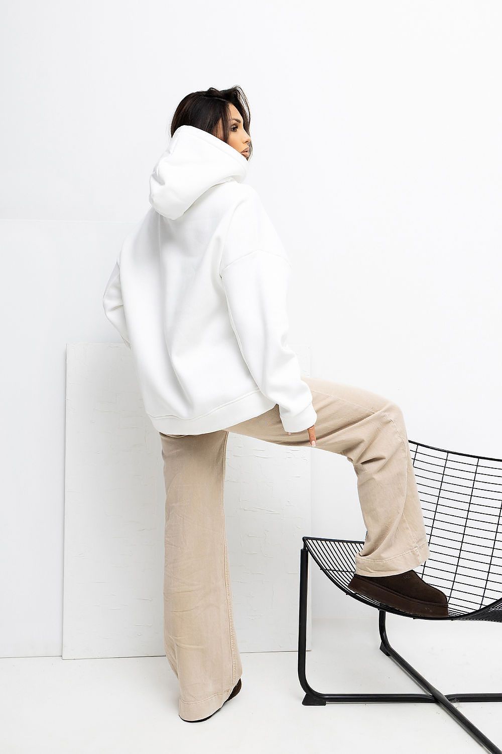 Person wearing a white hoodie and beige pants sitting on a black metal bench against a white wall.
