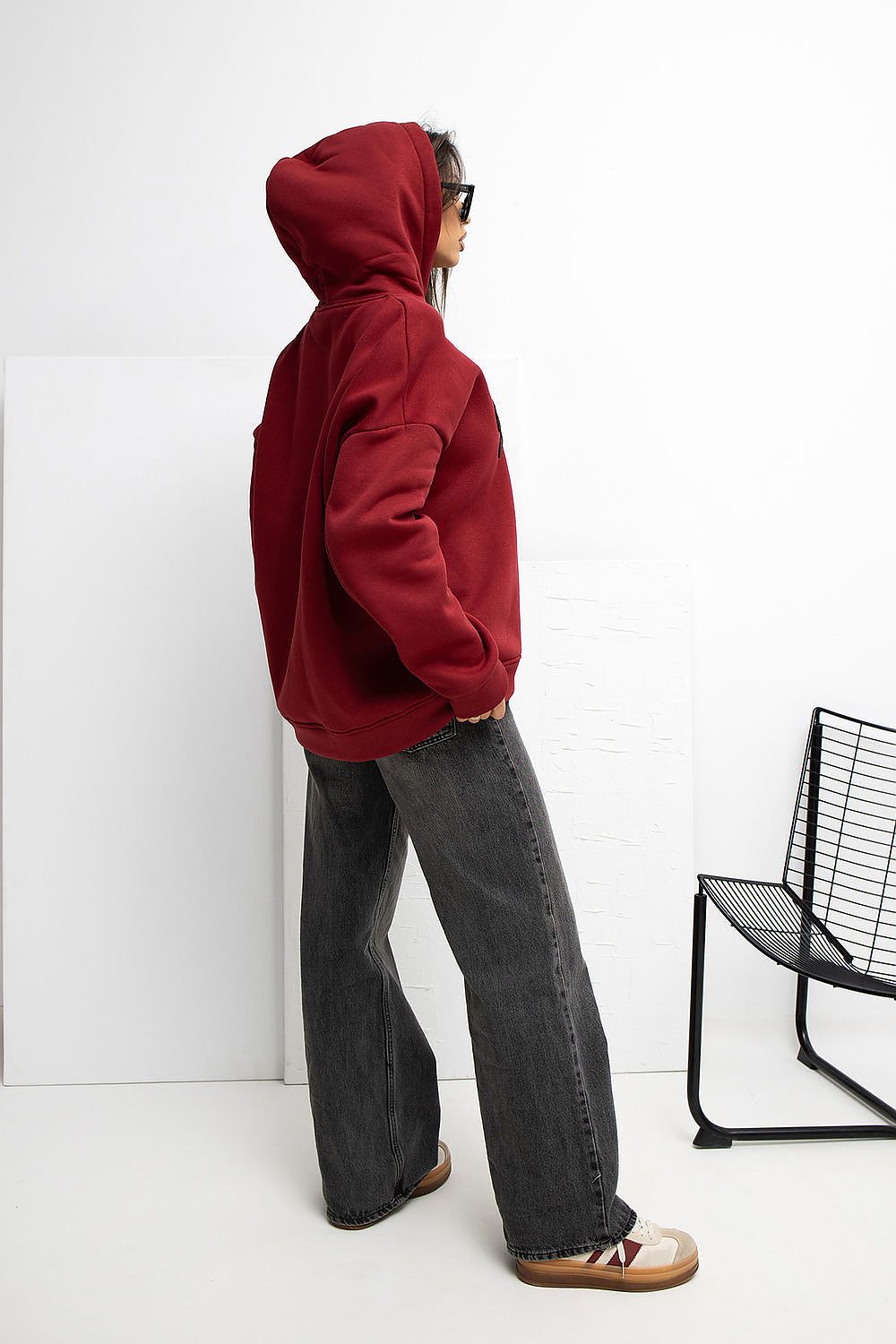 Person wearing a red hoodie and gray pants standing against a white wall with a black chair.