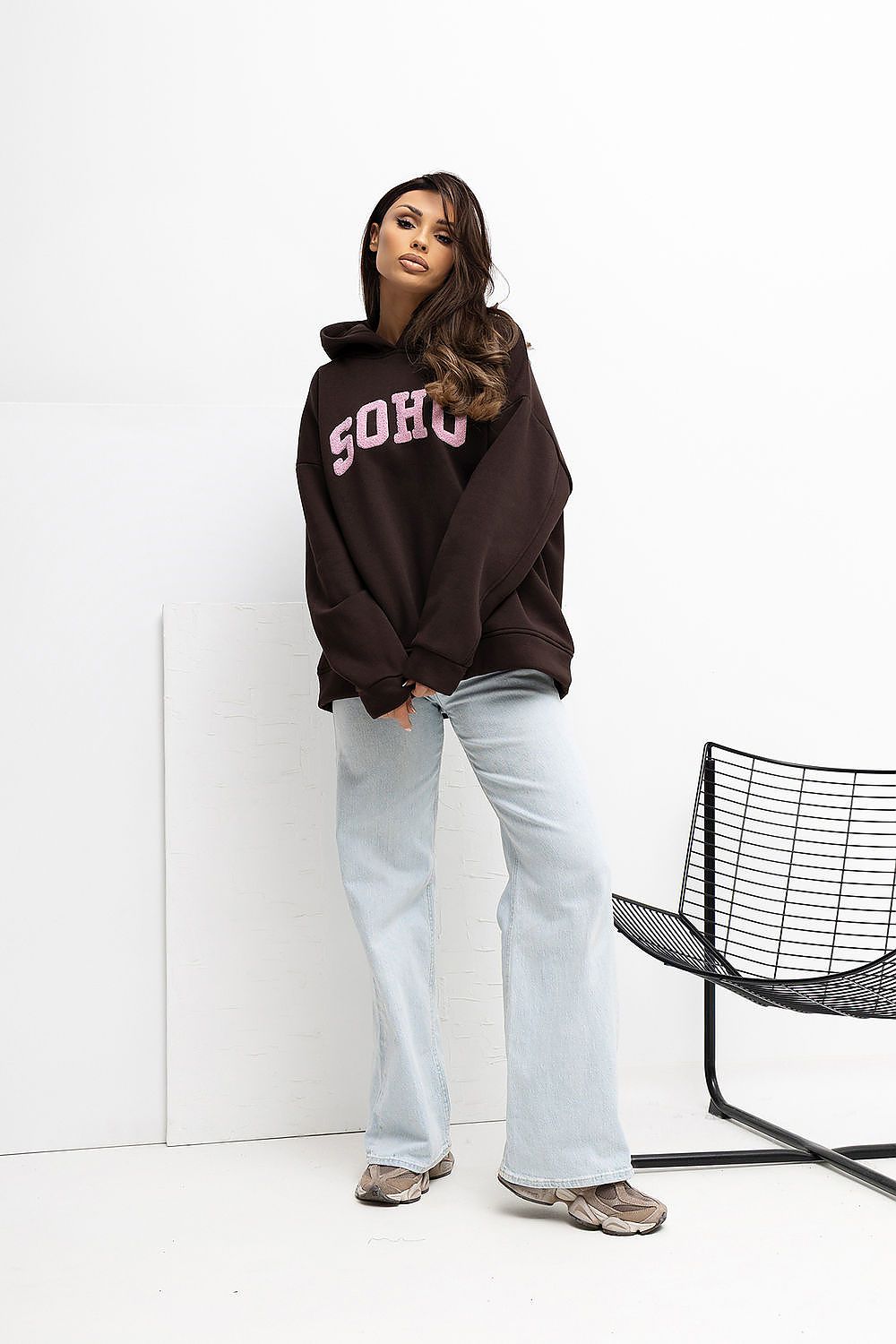Person wearing a dark hoodie with 'Soho' text, light blue jeans, and brown shoes on a white background.