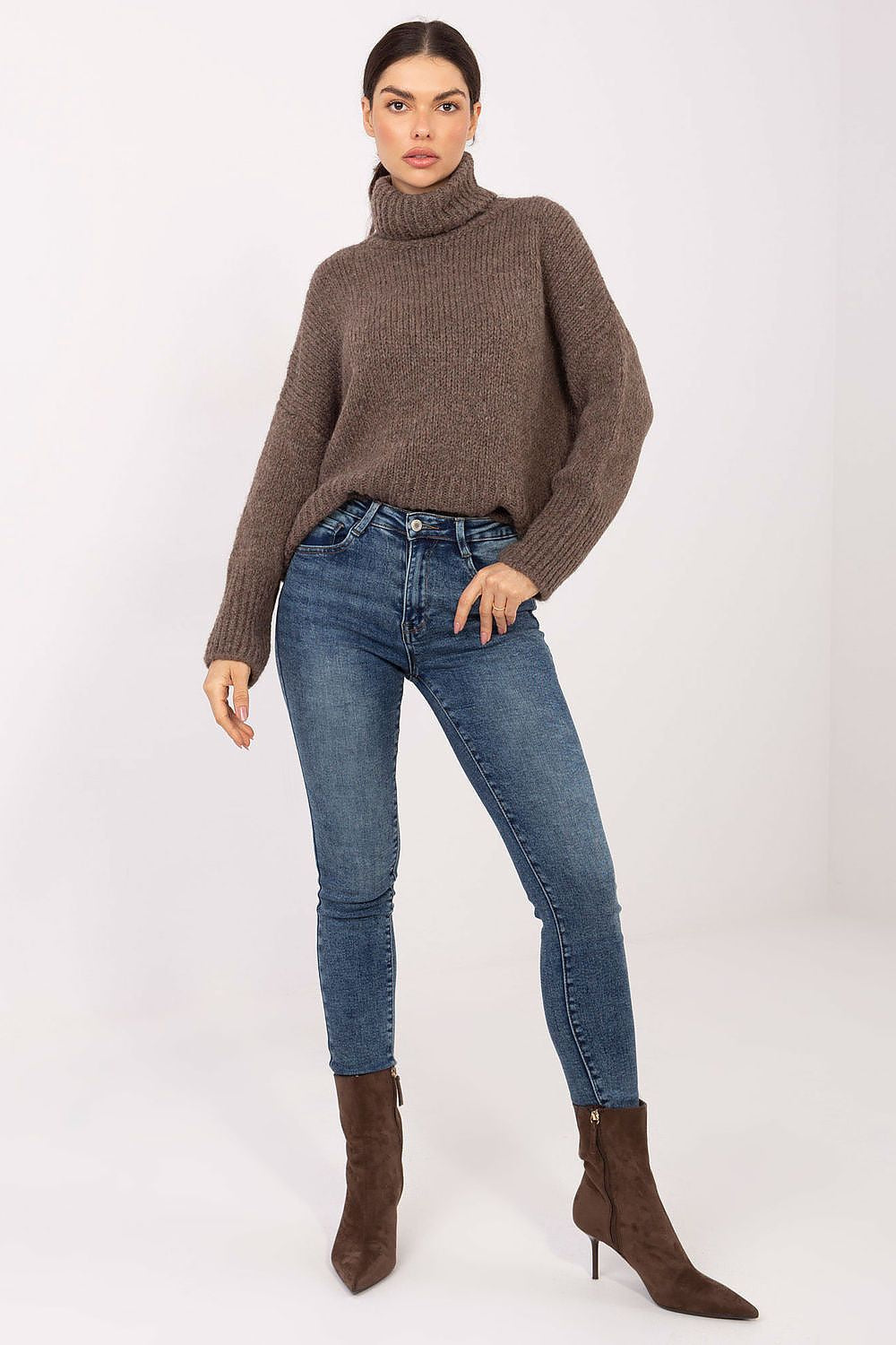 Woman wearing a brown turtleneck sweater, blue jeans, and brown boots on a white background
