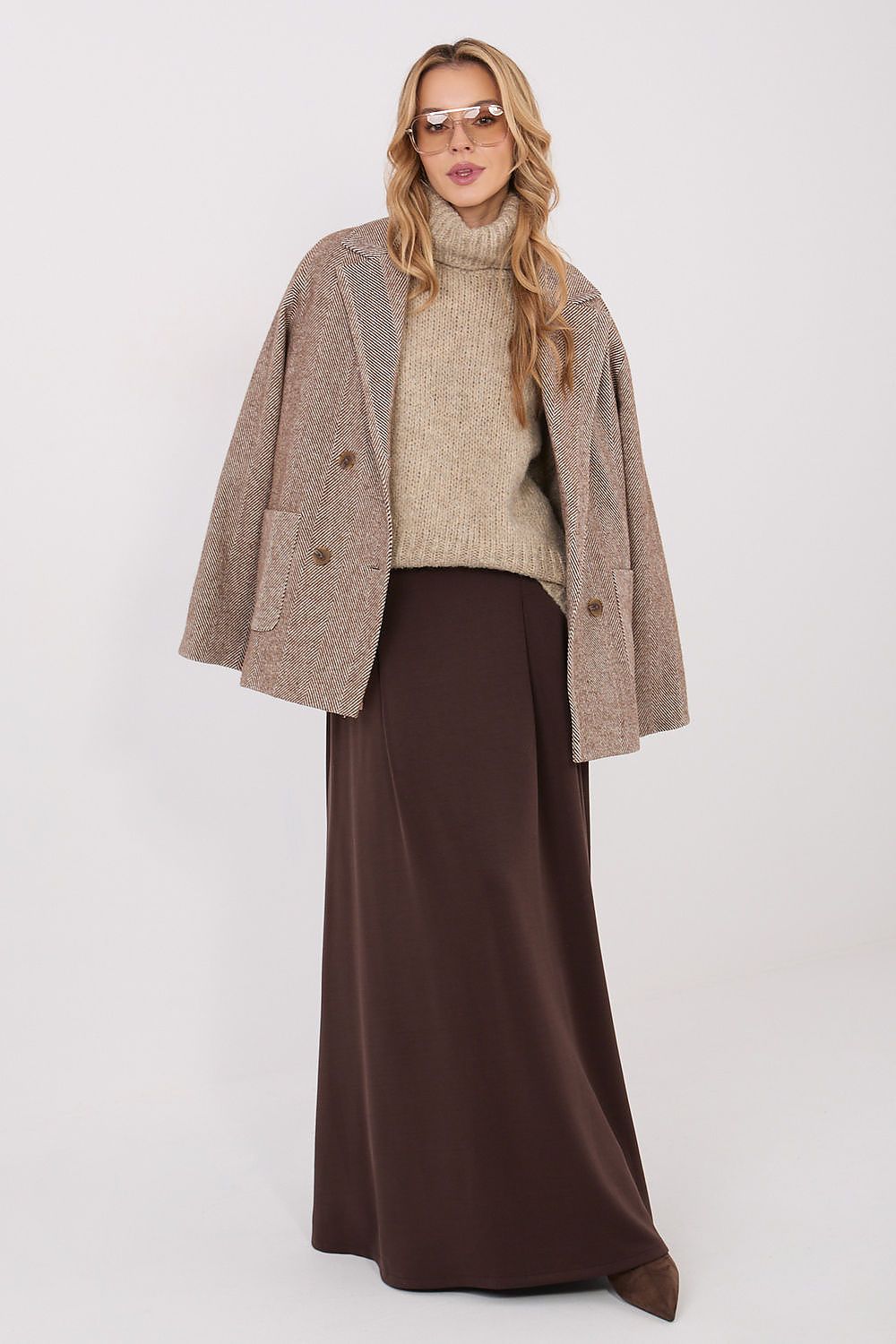 Woman wearing a beige sweater, brown coat, and long brown skirt on a white background
