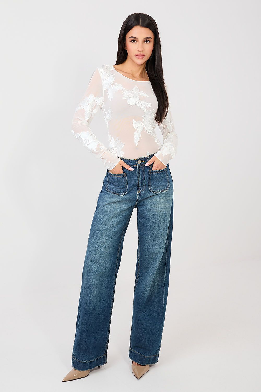 Woman wearing a white blouse with floral patterns and blue jeans on a light gray background